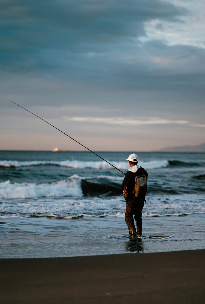 夕暮れの砂浜で、波打ち際から竿を振る釣り人。